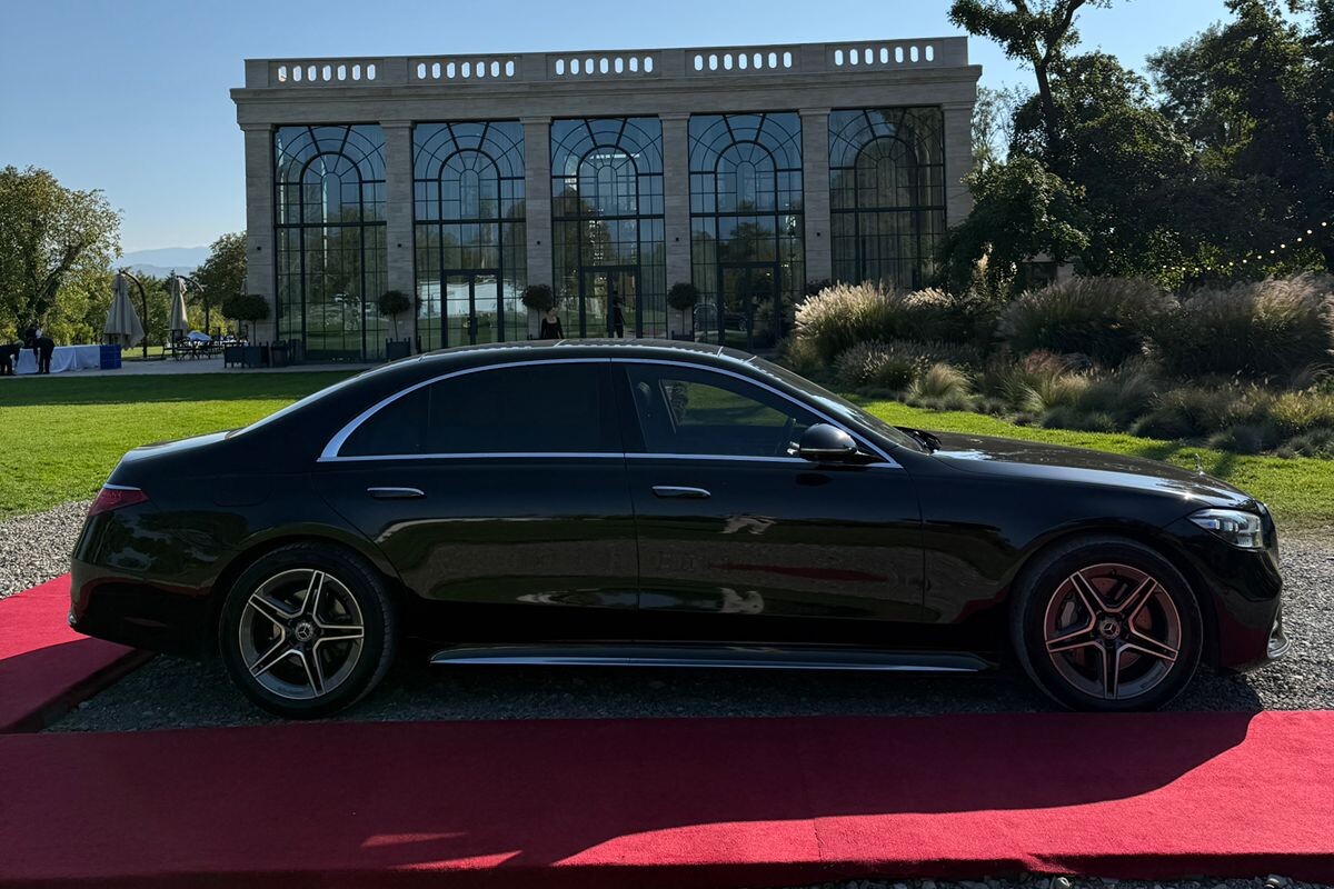 A black Mercedes S-Class waiting on a red carpet outside a glasshouse venue near Tbilisi, tall arched windows behind it.