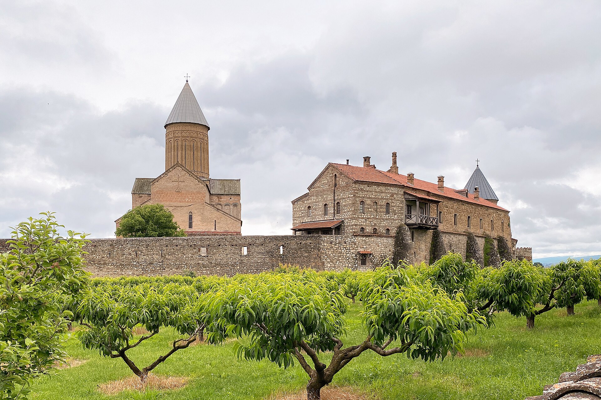 Alaverdi Cathedral rising above an orchard in the Alazani valley, its 11th-century stone walls and conical dome silhouetted against an overcast sky.