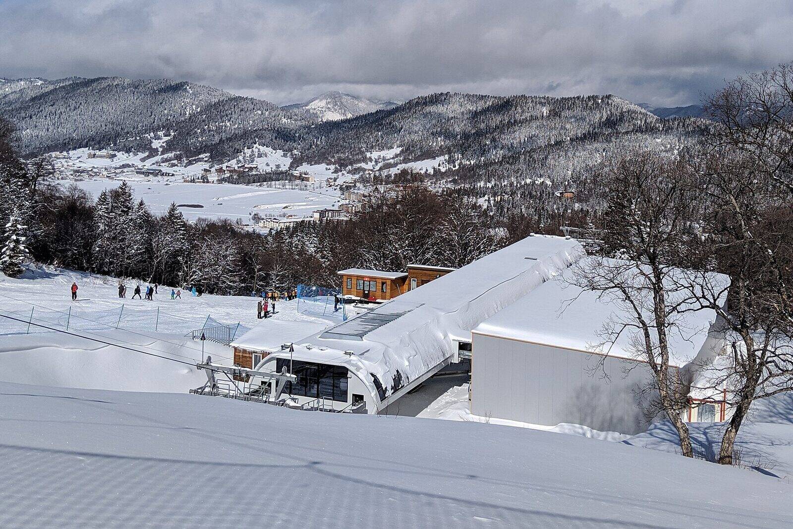 Fresh snow on a ski slope above Bakuriani, a lift station in the foreground and forested ridges beyond.