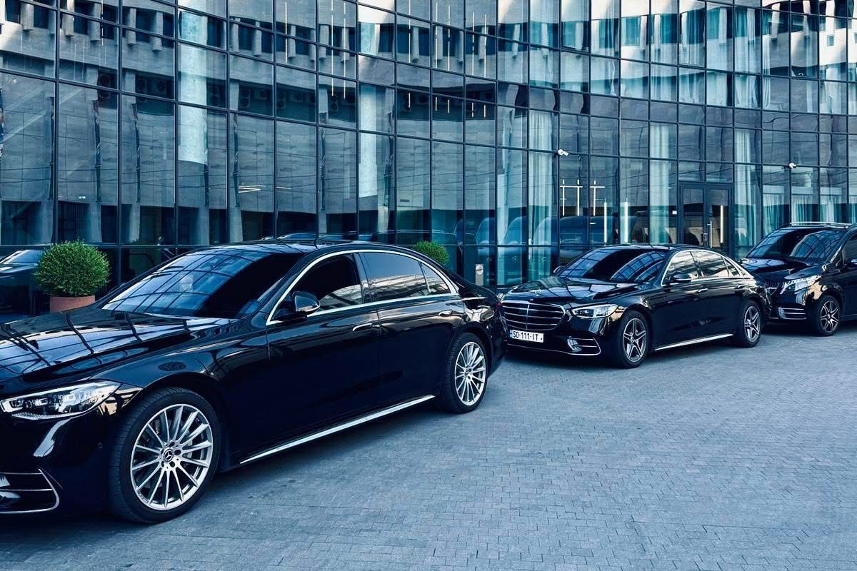 Three black Mercedes-Benz saloons lined up outside a curved glass office building on a bright day.