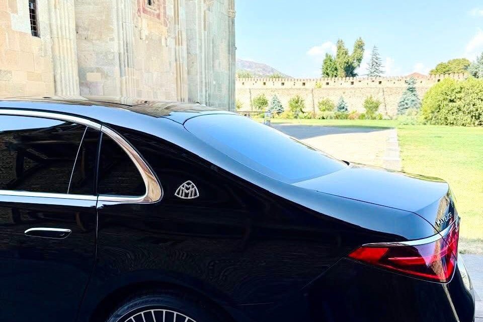 The rear of a black Mercedes-Maybach S-Class from the SOIT fleet beside the stone wall of Svetitskhoveli Cathedral in Mtskheta, the old fortress wall and lawn behind it.
