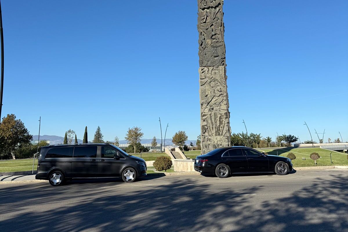 A black Mercedes-Benz V-Class and S-Class parked beside the Chronicle of Georgia monument in Tbilisi under a clear sky.