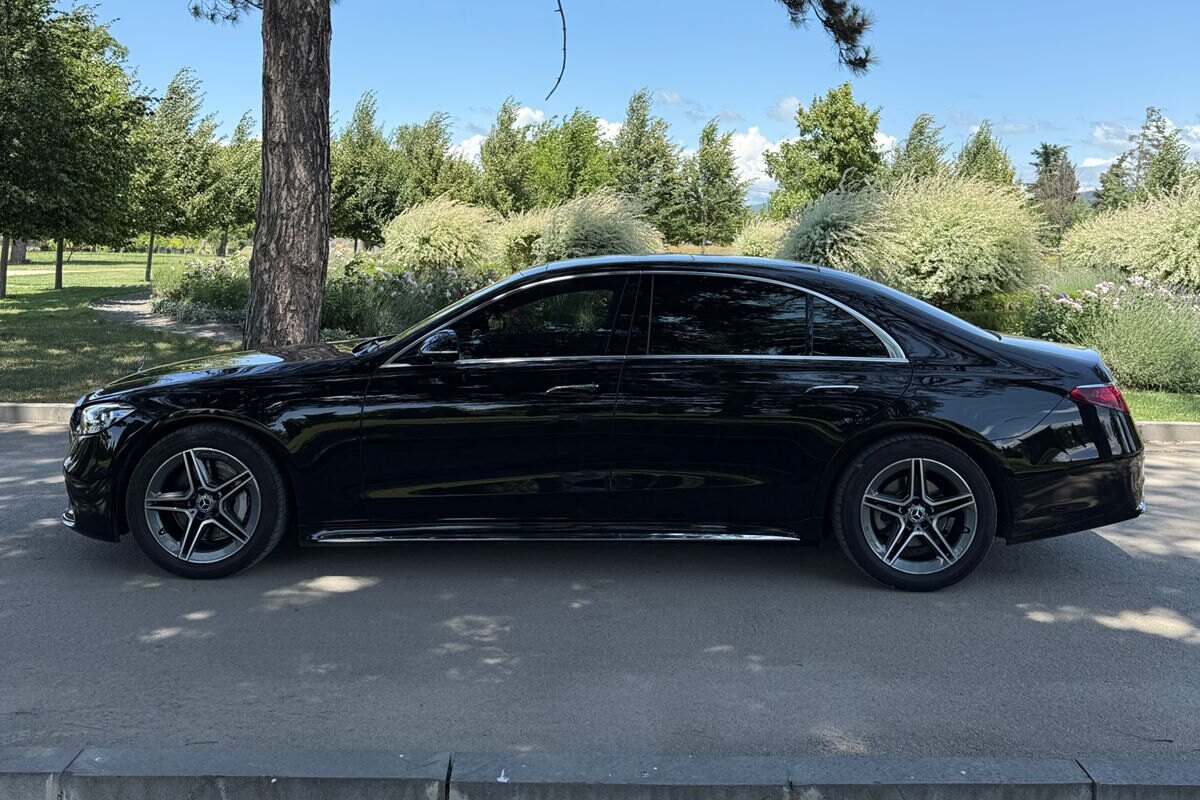 A black Mercedes S-Class from the SOIT fleet in side profile on a park road, trees and flowering shrubs behind it in summer.