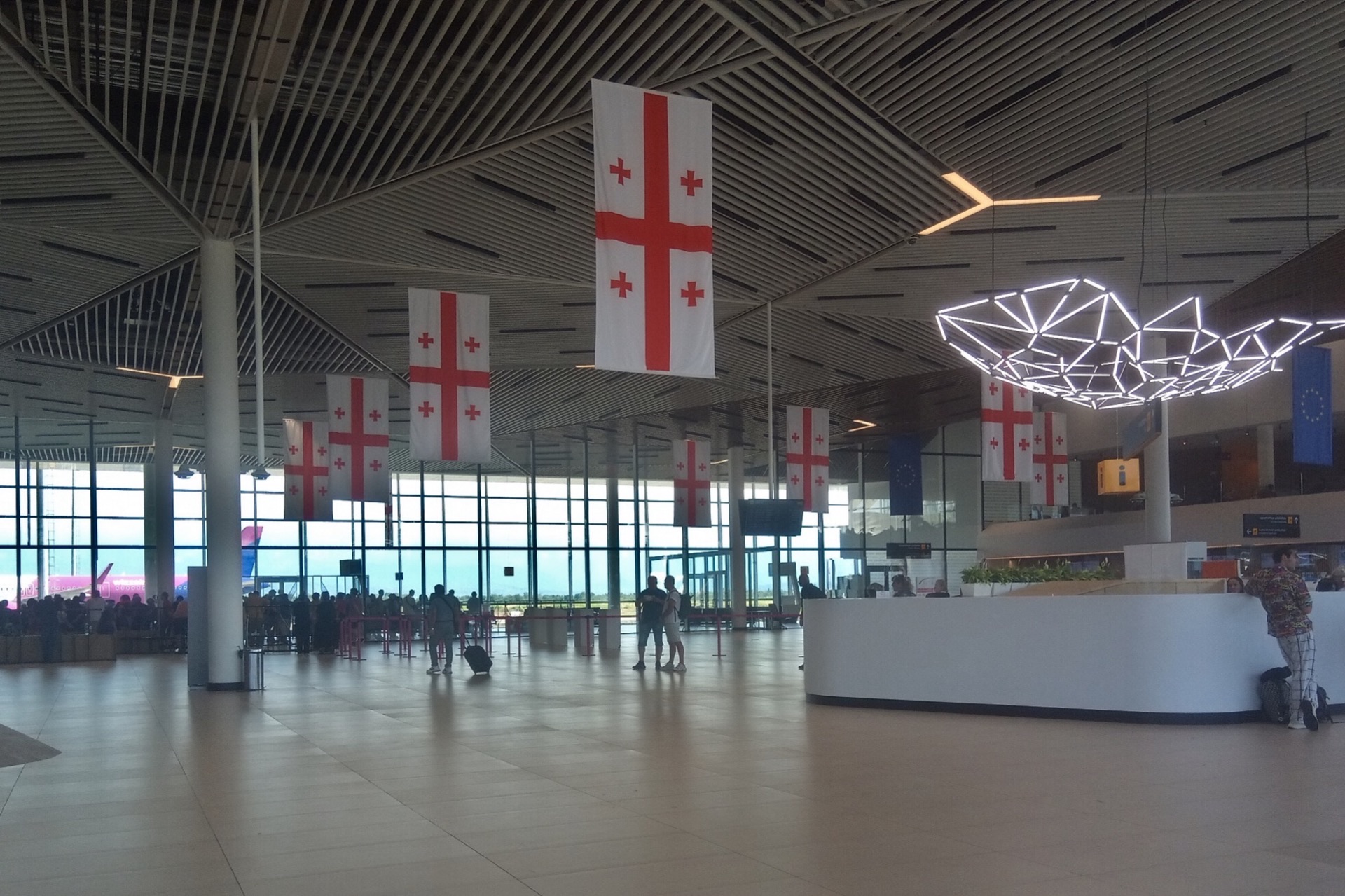 The departures hall at Kutaisi International Airport, Georgian flags hanging from the slatted ceiling and a low-cost aircraft visible through the glass.