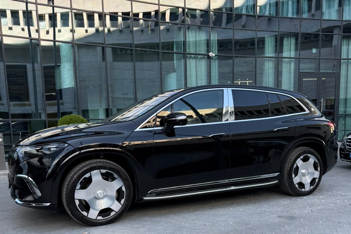 A black Mercedes-Maybach EQS 680 seen from the side, parked on paving outside a glass-fronted building in Tbilisi.