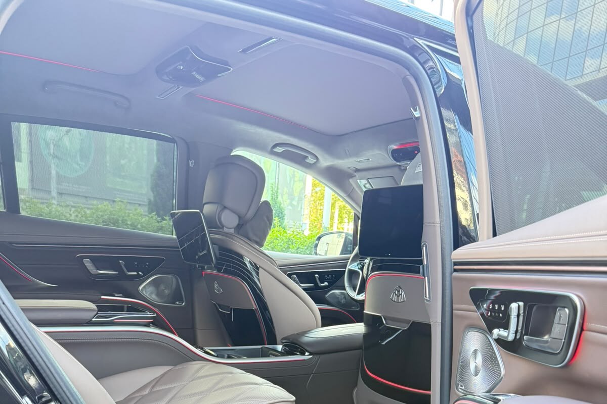 The rear cabin of the Mercedes-Maybach EQS 680 with the rear door open, showing two individual seats in light quilted leather and the illuminated MAYBACH sill plate.