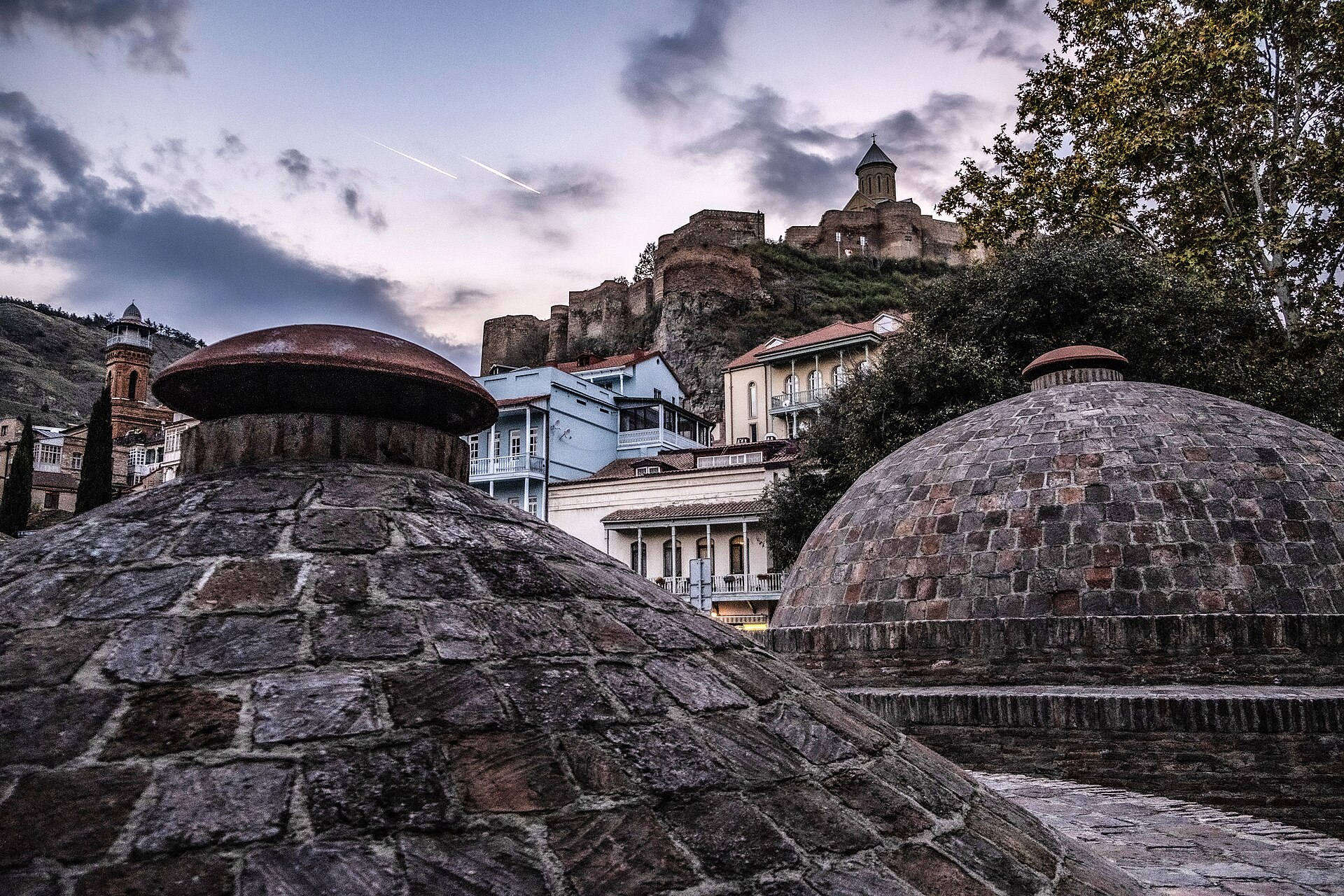 The brick beehive domes of the Abanotubani sulfur bathhouses in Tbilisi, the Narikala fortress walls rising on the hill behind them.