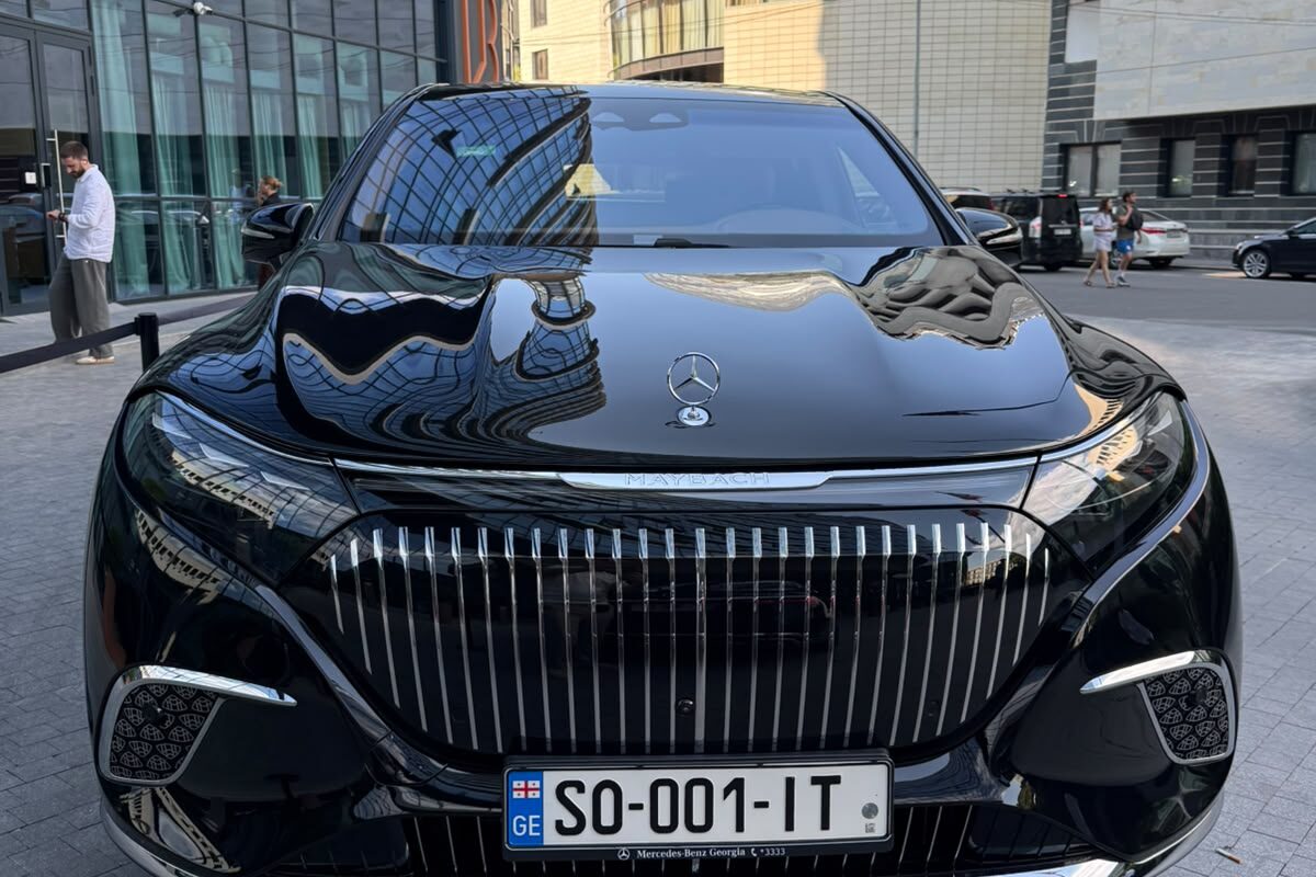 The front of a black Mercedes-Maybach EQS 680 on Georgian plates, parked on paving in Tbilisi with office buildings behind it.