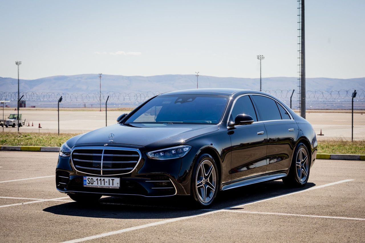 A black Mercedes-Benz S-Class waiting at the edge of a Georgian airfield, the Caucasus foothills visible in the distance.