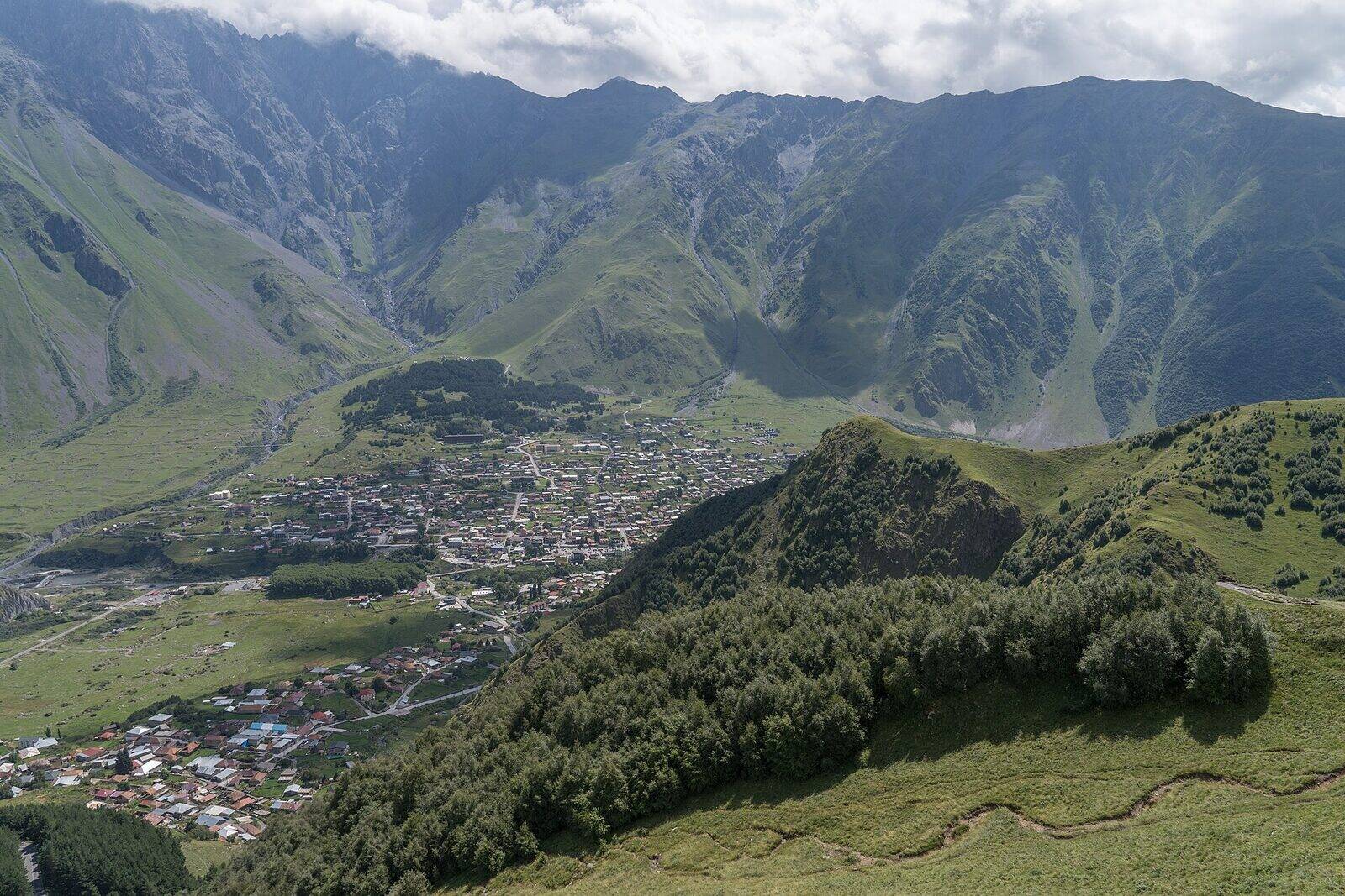 Stepantsminda spread along the valley floor below green Caucasus ridges, seen from the slope near Gergeti Trinity Church.