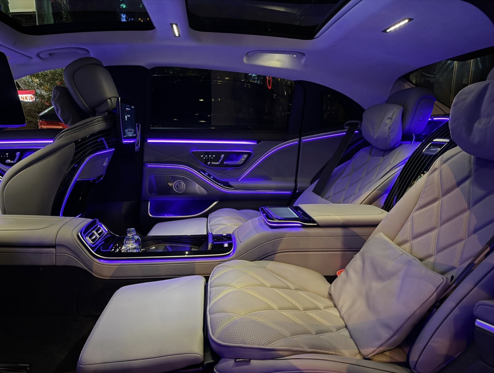 Interior of a Mercedes-Maybach rear cabin: cream leather diamond-stitched seats, purple ambient lighting, central console.