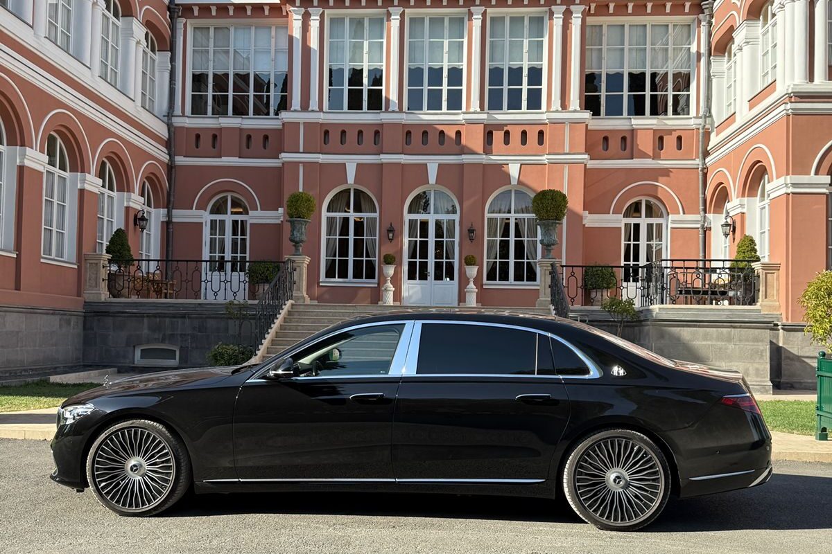 A black Mercedes-Maybach S-Class parked broadside in front of a red-brick mansion with arched windows and a wide stone staircase.