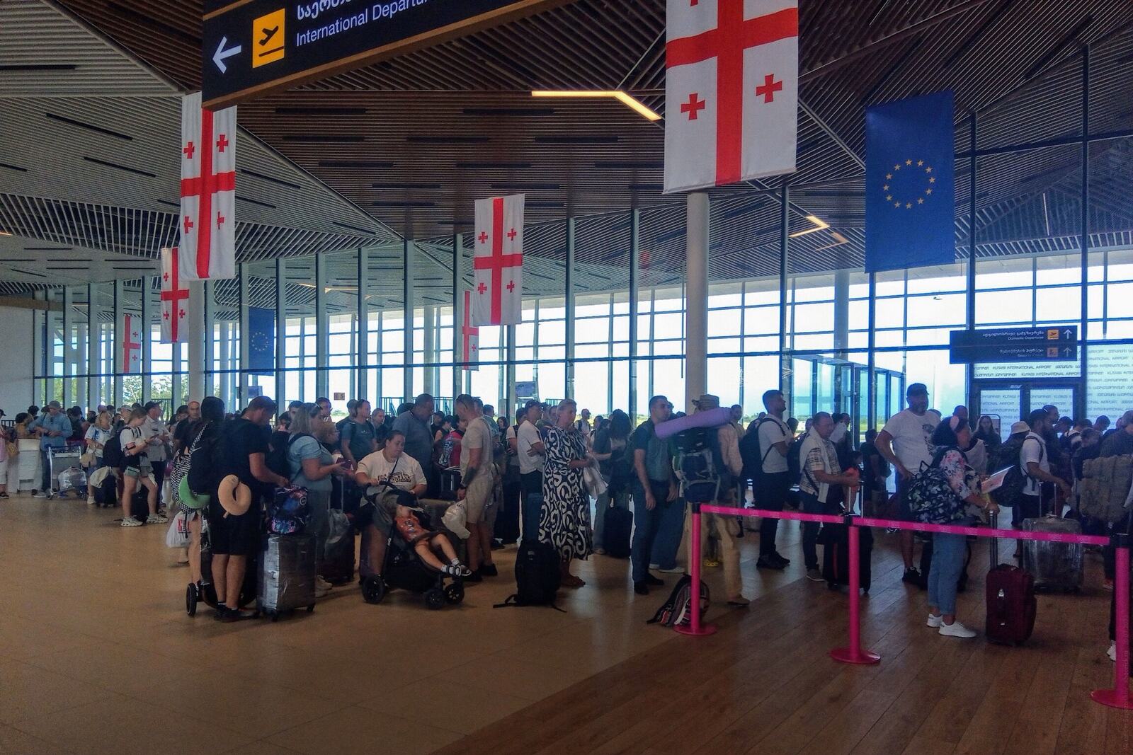 Passengers queuing at check-in inside the Kutaisi International Airport terminal, luggage trolleys and a low ceiling of pale slats.